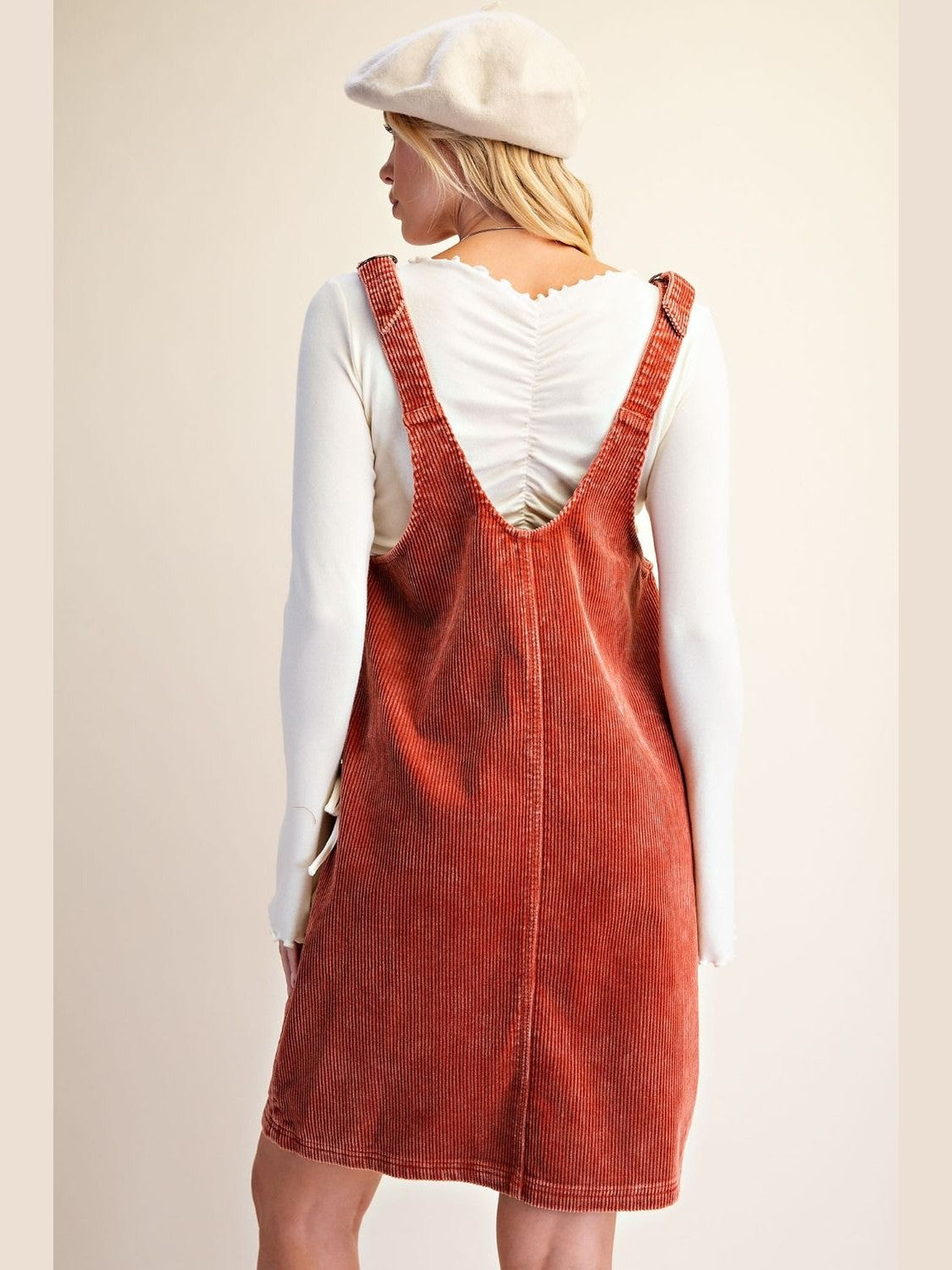 A woman shown from the back wearing rust-orange corduroy pinafore overalls over a white long-sleeve top, with a cream beret, against a plain beige studio background.