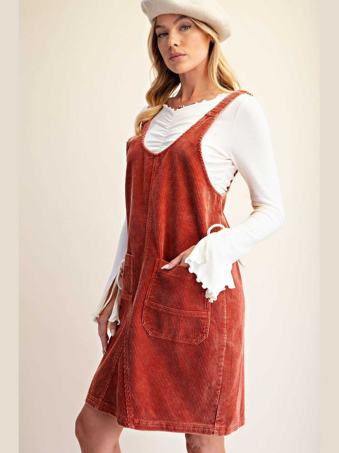 A woman models a rust-orange corduroy pinafore dress over a white long-sleeve ruffled shirt, wearing a matching white beret against a warm beige studio background.