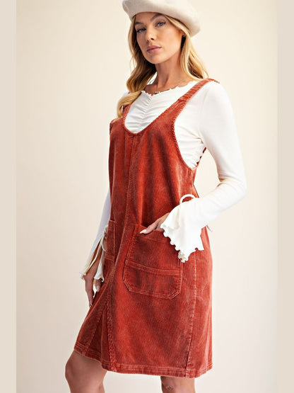 A woman models a rust-orange corduroy pinafore dress over a white long-sleeve ruffled shirt, wearing a matching white beret against a warm beige studio background.