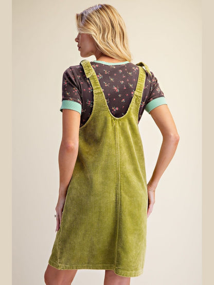 A woman shown from the back wearing an olive green corduroy pinafore dress over a short-sleeve floral t-shirt with mint green cuffs, against a solid beige background.