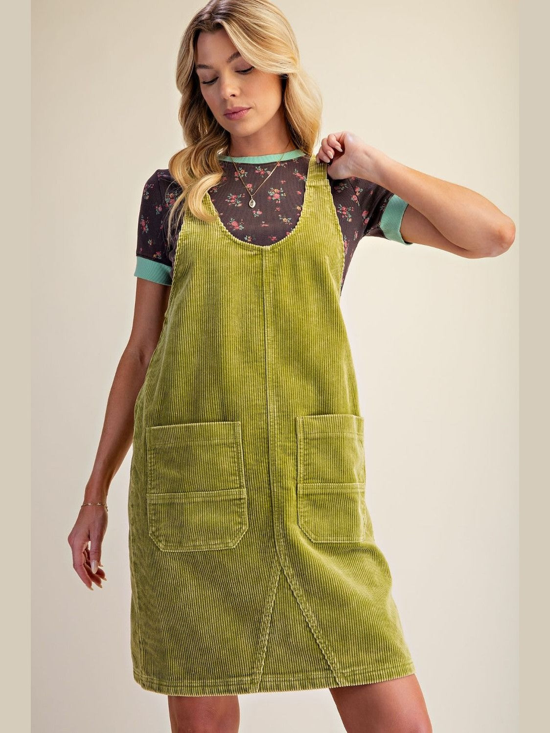 A woman modeling a sage green corduroy pinafore dress over a short-sleeve floral t-shirt, set against a plain beige studio background.