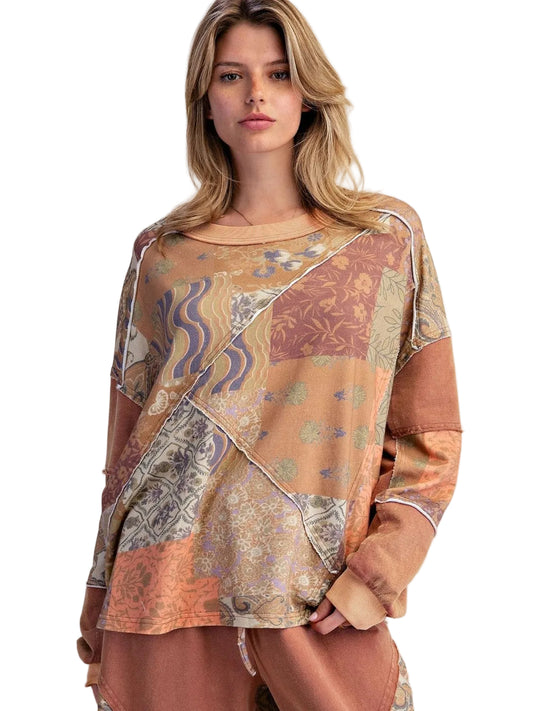 A woman modeling a loose, long-sleeve top with a patchwork-style abstract print in warm earthy tones of beige, brown, orange, and muted blue, against a plain light beige studio background.