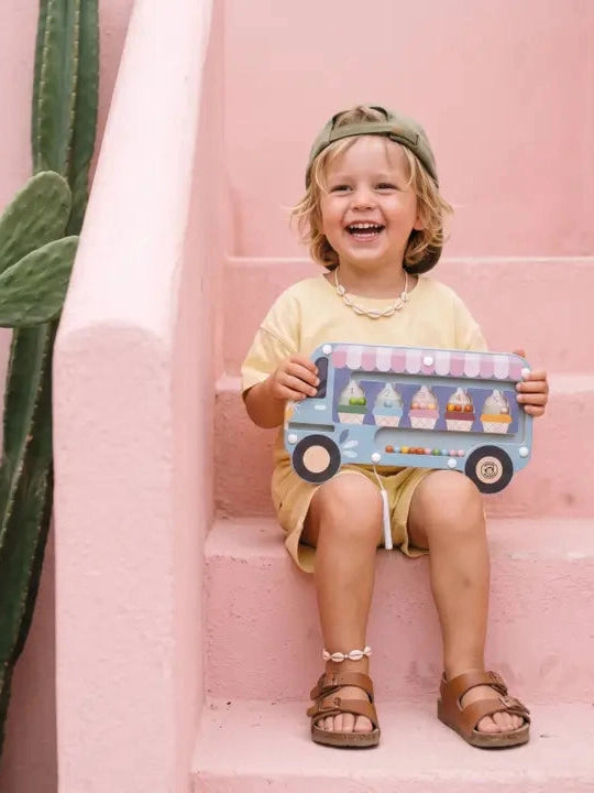 Child holding a toy ice cream truck against a pink wall with a cactus plant.