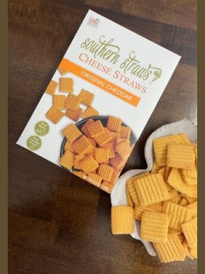 Box of southern strands cheese straws on a wooden surface with cheese straws beside it.
